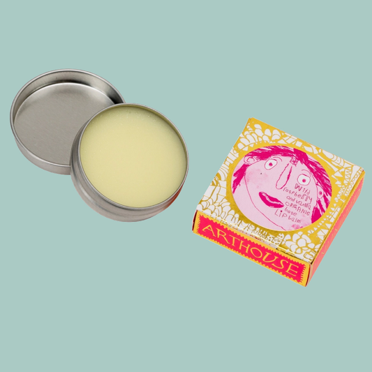 An open round metal tin of balm with pale yellow balm visible inside, shown beside its illustrated retail box. The box is yellow, pink, and white with a cartoon face and the brand name ARTHOUSE visible.