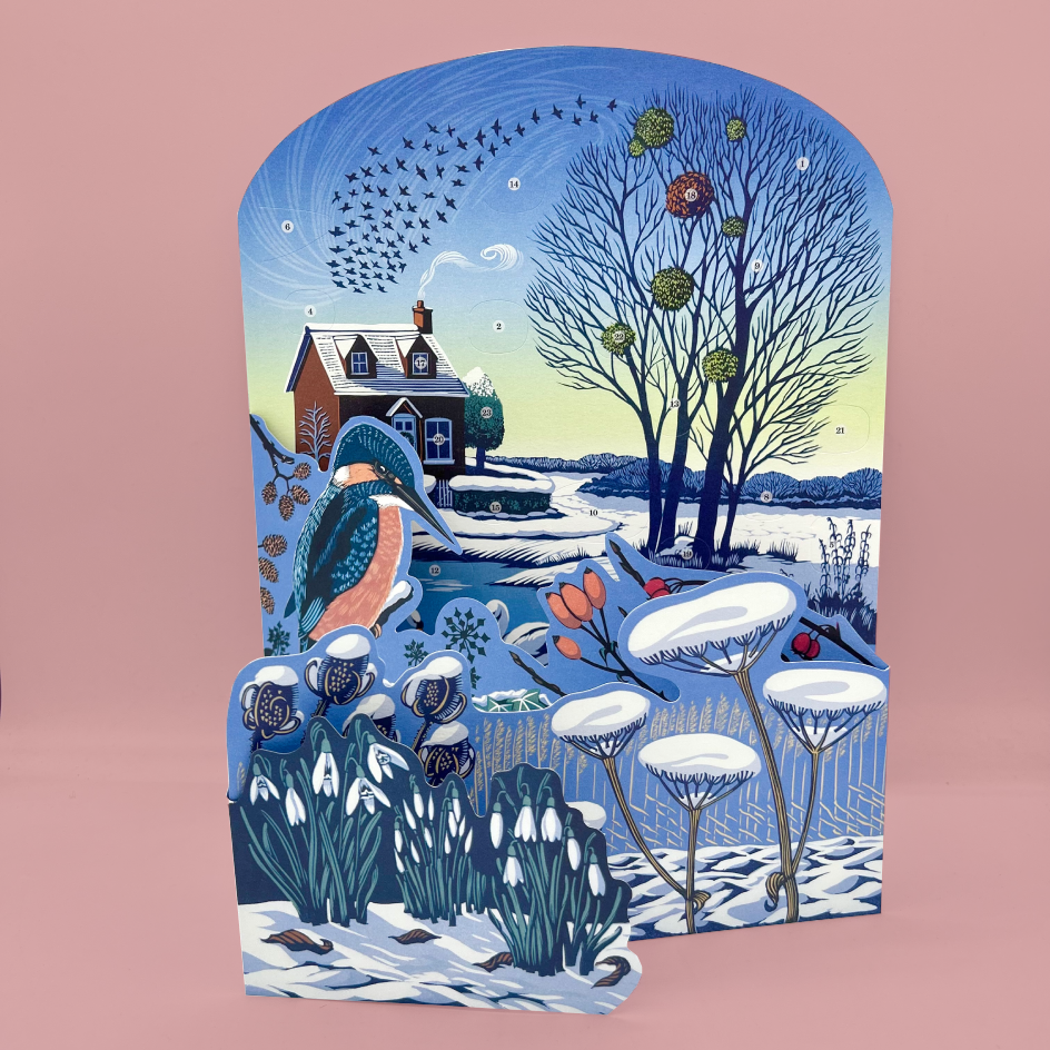 A decorative winter concertina advent calendar with an illustrated snowy cottage, bare trees, birds, a bluebird, and snow-covered plants, shown against a solid pink background.