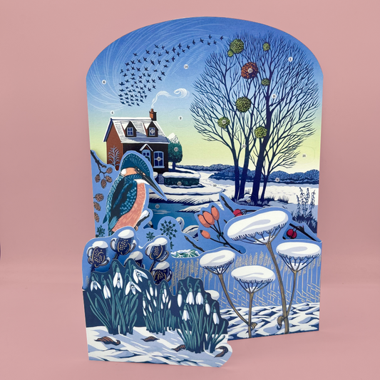 A decorative winter concertina advent calendar with an illustrated snowy cottage, bare trees, birds, a bluebird, and snow-covered plants, shown against a solid pink background.