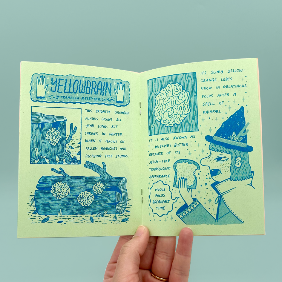 A hand holding an open illustrated zine with blue drawings and text on pale yellow-green paper. The pages show a whimsical nature-themed layout with the title 'Yellowbrain' and illustrated elements including a brain, tree logs, and a stylized face wearing a witch hat.