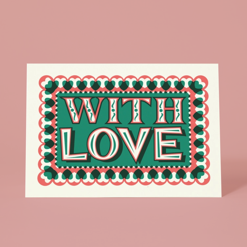 A  greeting card with a retro-style graphic design that reads 'WITH LOVE' in bold white letters with red and dark green outlines, set against a teal background. The card is framed by a scalloped border of red and dark green hearts, and it is photographed on a solid dusty pink background.
