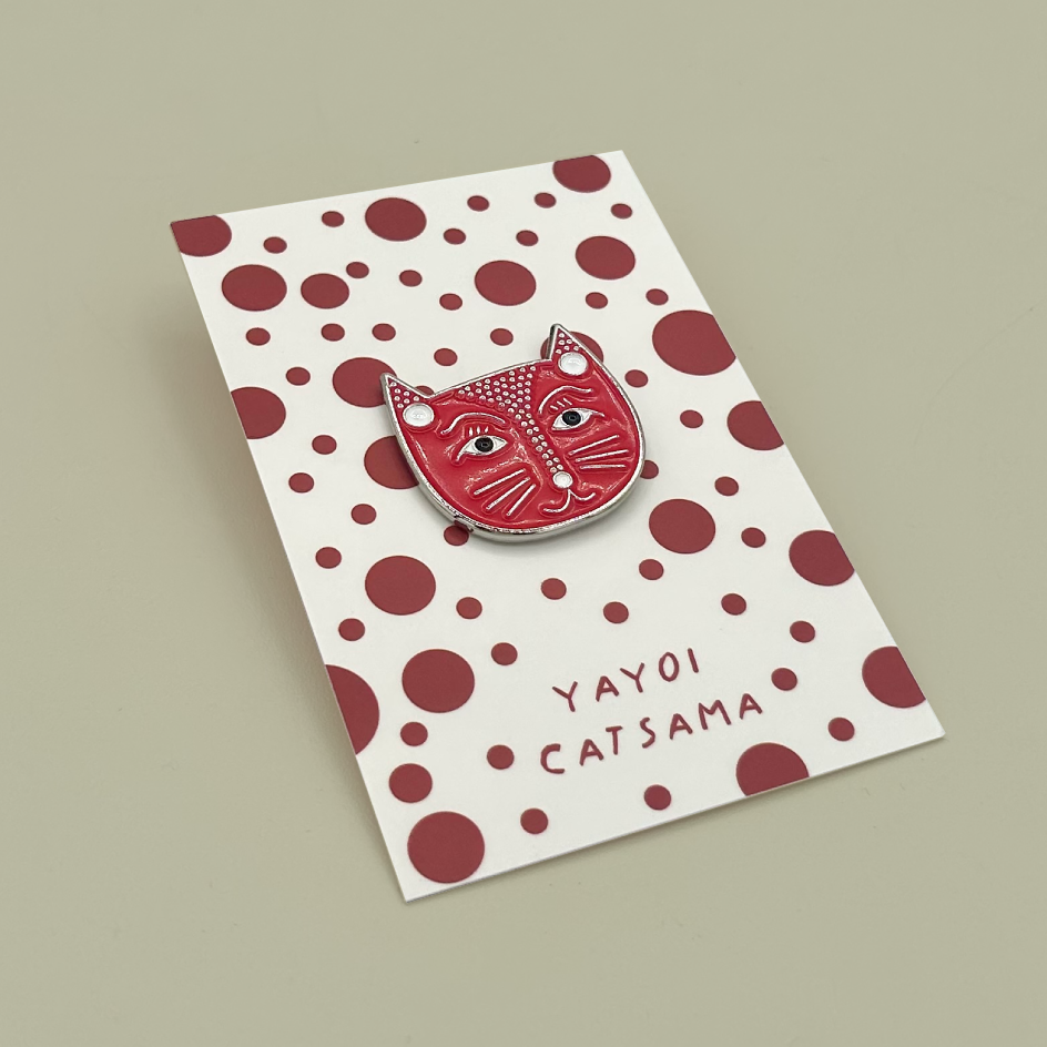 A red and white cat-face enamel pin with white polka dots and the text 'YAYOI CATSAMA' on a white backing card with red polka dots, photographed on a plain beige background.