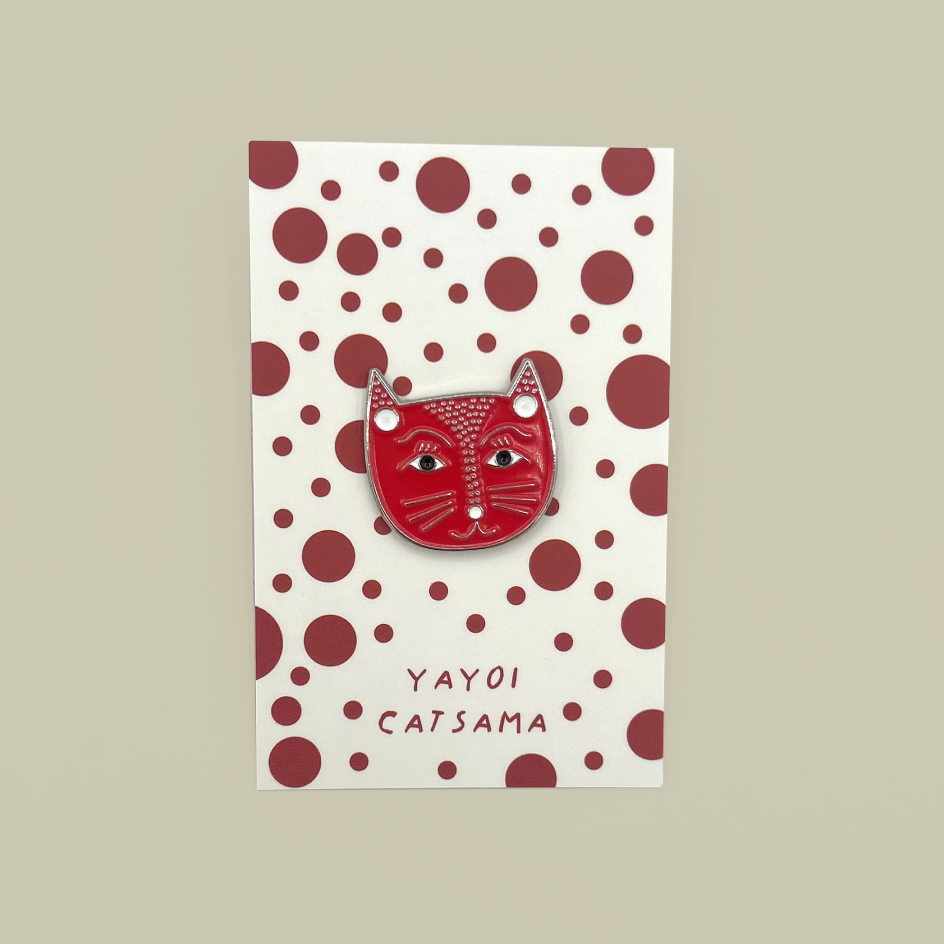A red cat-face enamel pin with white and black details, mounted on a cream card with dark red polka dots and the text 'YAYOI CATSAMA'.
