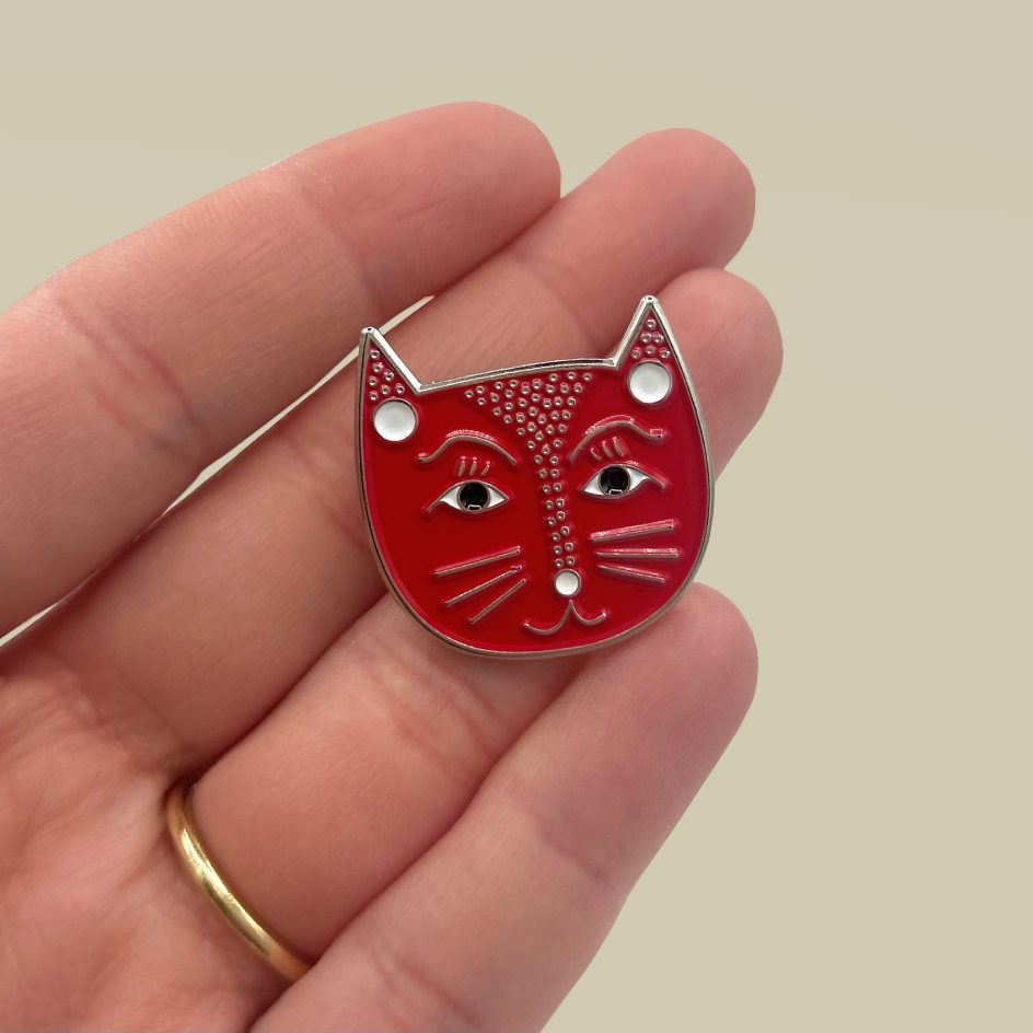 A close-up of a hand holding a small red cat-face enamel pin with silver metal outlines and white dot details, against a plain warm beige background.