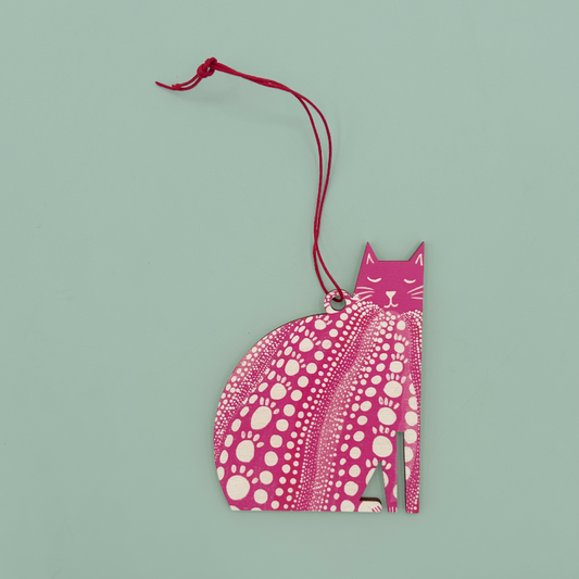 A pink cat-shaped ornament with a red hanging string, featuring a white polka dot pattern on the body and a solid pink head, set against a solid light green background.