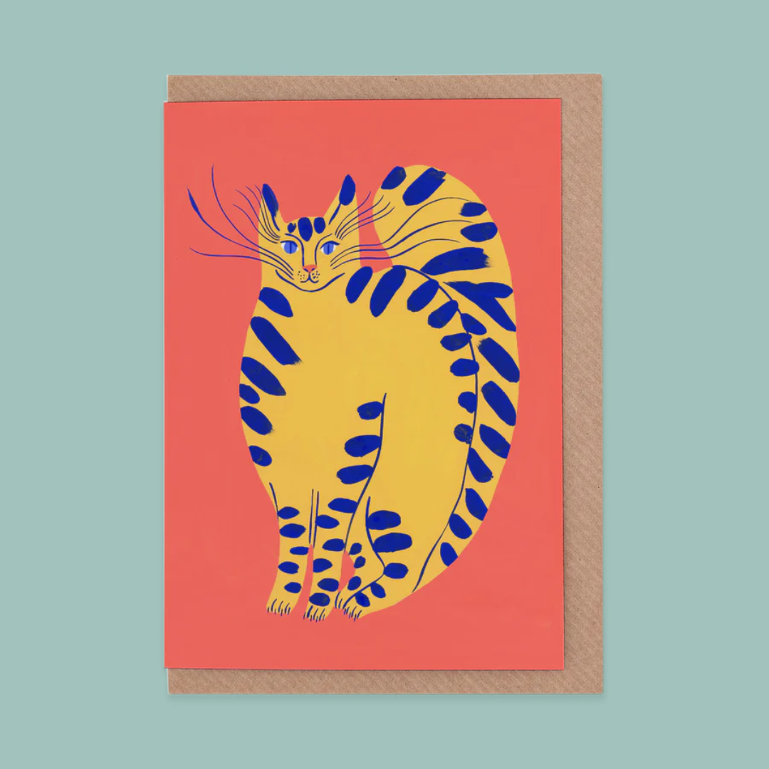 Greeting card with a yellow cat illustration on a coral coloured background. The card is on a teal backdrop.