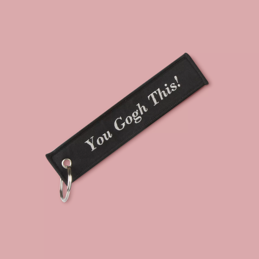 A black fabric keychain with white embroidered text reading 'You Gogh This!' and a silver metal carabiner, shown on a solid pink background.