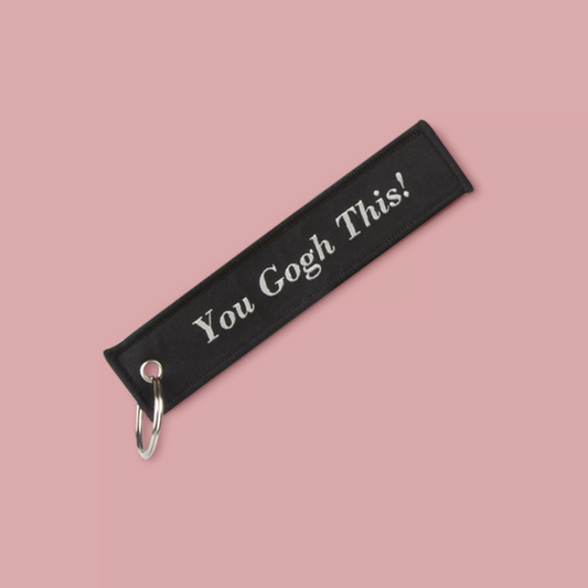 A black fabric keychain with white embroidered text reading 'You Gogh This!' and a silver metal carabiner, shown on a solid pink background.
