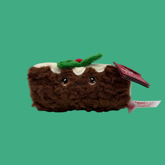 A brown plush toy shaped like a slice of cake or pudding, with white icing, a green holly accent, and embroidered eyes. A red hangtag is attached on the right side, photographed on a clean light gray studio background.